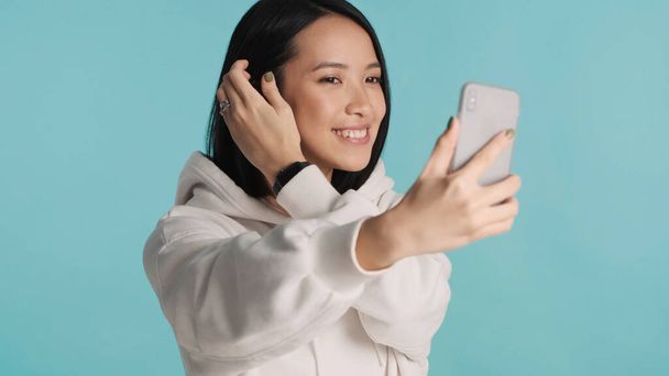 Ασιάτισσα που φοράει κουκούλα και βγάζει selfie στο smartphone πάνω από πολύχρωμο φόντο. Σύγχρονη τεχνολογία έννοια. Όμορφη γυναίκα που ποζάρει στο στούντιο - Φωτογραφία, εικόνα