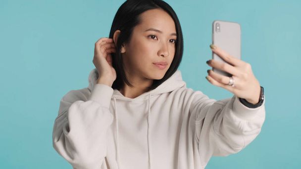 Azjatycka dziewczyna wygląda pewnie trzymając smartfona robiąc selfie na kolorowe tło. Milenijny twórca treści - Zdjęcie, obraz