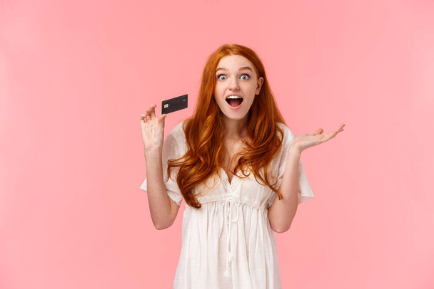 Waist-up portrait amused, wondered and excited fashionable feminine redhead girl telling about great promos and bonuses in bank, holding credit card and smiling astonished, pink background. - Photo, Image
