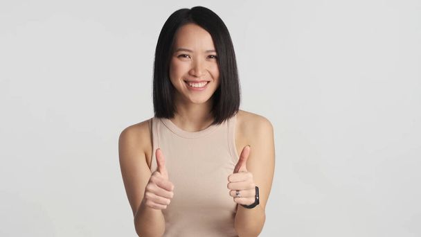 Iloinen aasialainen nainen pitää peukalot erillään valkoisella pohjalla. Aasialainen nainen näyttää onnelliselta hyväksyessään hyvän idean. Kuten se ele - Valokuva, kuva