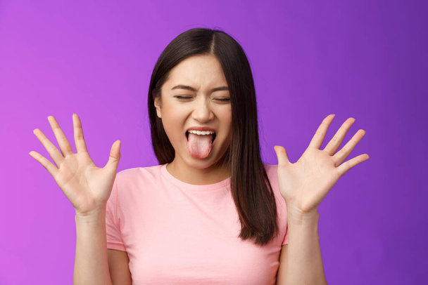 Close-up silly moody asian girl complaining act immature and bothered, raise hands unsatisfied show tongue aversion disgust, close eyes, react terrible disgusting thing, stand purple background. - 写真・画像