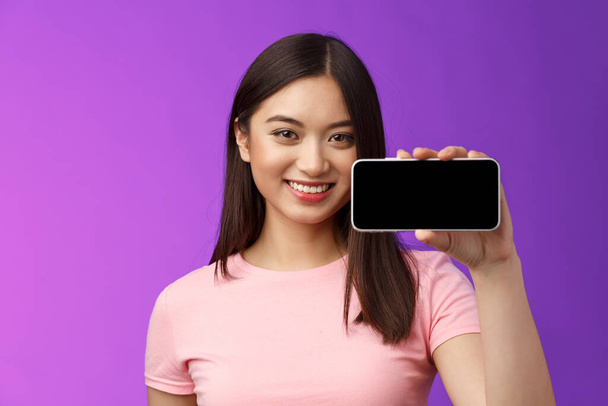 Κοντινό πλάνο χαρούμενη χαριτωμένο ασιατική γυναίκα κατέχουν smartphone οριζόντια, δείχνουν οθόνη του τηλεφώνου, να εισαγάγει φοβερό νέα εφαρμογή, παίζοντας ενδιαφέρον τηλεφωνικό παιχνίδι, να σταθεί μωβ φόντο προώθηση. - Φωτογραφία, εικόνα