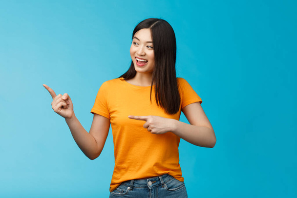 Přátelsky nadšený asijské dívka zvědavě dívá stranou, ukazuje vlevo zajímavý objekt, šklebí radostně diskutovat o novém otevřeném obchodě, stojí modré pozadí bezstarostně živé mluvení. - Fotografie, Obrázek