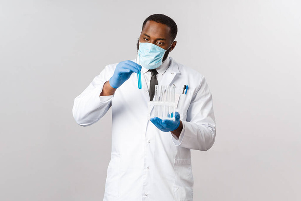 Ковид-19, лечение коронавируса и лабораторная концепция. Сосредоточенный и осторожный афро-американский врач в латексных перчатках и медицинской маске для лица, серьезный взгляд на пробирку, изобретение лекарства, вакцина
. - Фото, изображение