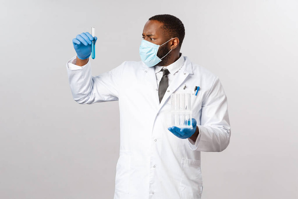 Covid-19, θεραπεία ασθενών με κορωνοϊό και εργαστηριακή αντίληψη. Σοβαρή εμφάνιση αφροαμερικανός γιατρός προσπαθεί να κάνει εμβόλιο, κορόνα φάρμακο, κοιτάζοντας test-tube με μπλε υγρό φάρμακο. - Φωτογραφία, εικόνα