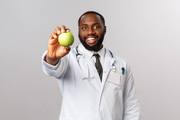 医療、医療、健康的なライフスタイルの概念。アップル1日。陽気でハンサムなアフリカ系アメリカ人の医師は、患者に緑のリンゴを与え、果物を示して果物やビタミン、灰色の背景を食べるように頼む. - 写真・画像