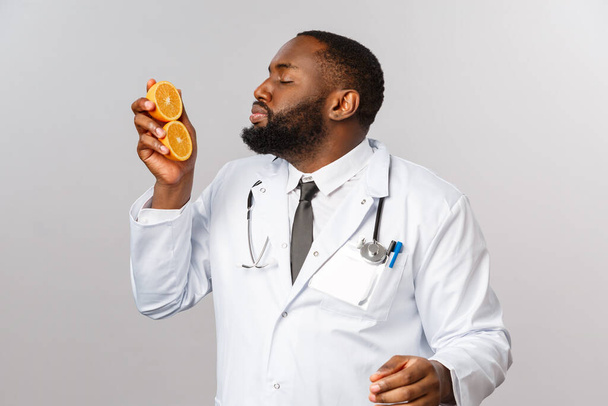Atención médica, dieta saludable y concepto de enfermedad. Apasionado divertido afroamericano médico olfatear vitamina c, como comer frutas, prescribir más naranjas y alimentos saludables, fondo gris
. - Foto, imagen