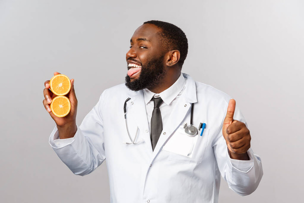Gezondheidszorg, gezonde voeding en ziektebegrip. Gelukkig grappig Afrikaans-Amerikaanse arts geven aanbeveling veilig te blijven, het eten van vitaminen in groenten en fruit, show thumb-up en likken oranje. - Foto, afbeelding