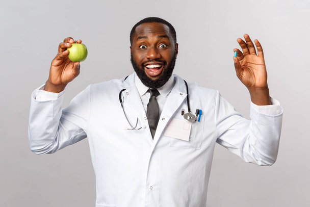 医療、医療、健康的なライフスタイルの概念。熱心なアフリカ系アメリカ人医師のアドバイスは、より多くのビタミン、果物、リンゴと錠剤を食べ、灰色の背景に立って健康にとどまる. - 写真・画像