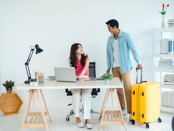 Happy Asian couple, young woman with laptop on table and man standing and holding suitcase succeed with trip planning together at home background, ready to travel, happy holiday and vacation concept. - Foto, afbeelding