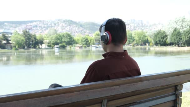 Disparo desde detrás del hombre sentado contra la vista de la orilla del río y escuchar música, tiempo para sentarse y relajarse con la vista, tiempo libre mientras escucha música por auriculares - Metraje, vídeo