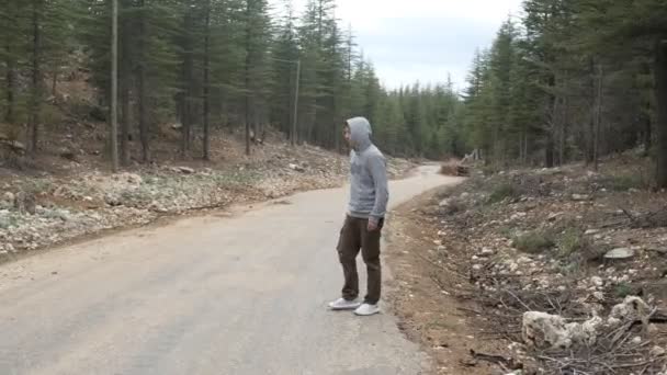 Ormanda asfalt yolda otostop çeken genç adam, ara sıra kapüşonlu adamın el hareketlerini durdur, dağ yolunda aracı durdur. - Video, Çekim