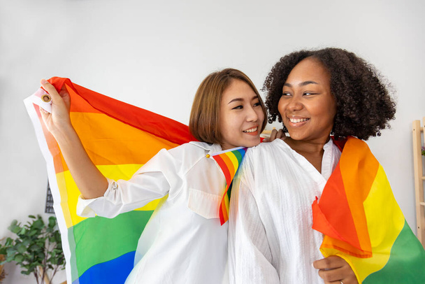 Couple of same sex marriage from difference races holding LGBTQ rainbow flag for pride month to promote equality and differences of homosexual and discrimination - Zdjęcie, obraz