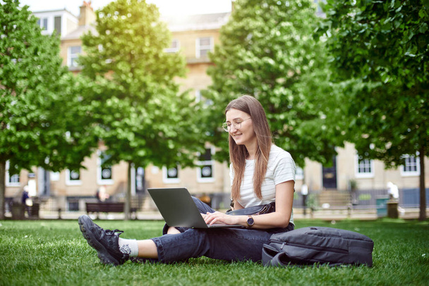 Молодая девушка улыбается, глядя на свой ноутбук. Женщина работает в ноутбуке, сидя на траве. Женщина проходит онлайн-курсы в парке, сидя на лужайке. Концепция удаленного офиса. - Фото, изображение