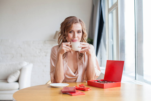 Happy young woman eating chocolate candies and drinking coffee - Foto, Imagen