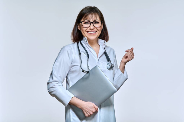 Portrait of female doctor in white uniform with laptop stethoscope, confident woman looking at camera, on light studio background. Technology online consultation service, medicine health care concept - Foto, immagini