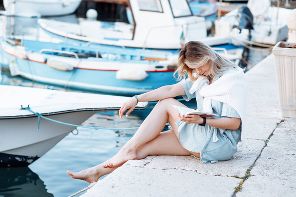 Thoughtful barefoot blond woman reading book near boats, river port or dock outdoors. Sunny day on vacation or weekend, relaxation period. Education, self preparing, learning new. Bookworm on travel - Photo, image