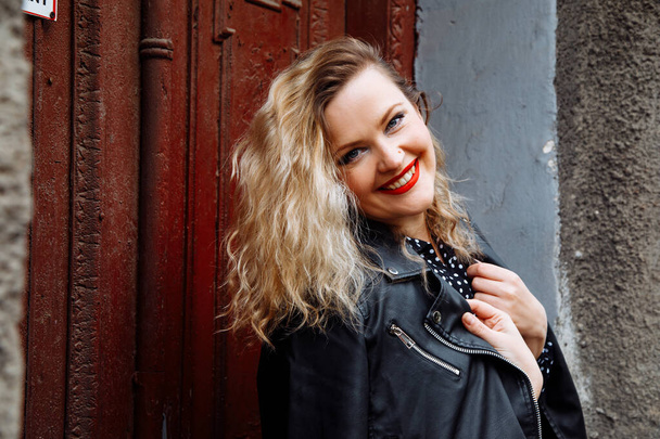Modern smiling stylish blond woman with make-up posing, looking at camera in cool black leather jacket and dress against brown house door. City street style. Having friendly conversation and have fun - 写真・画像