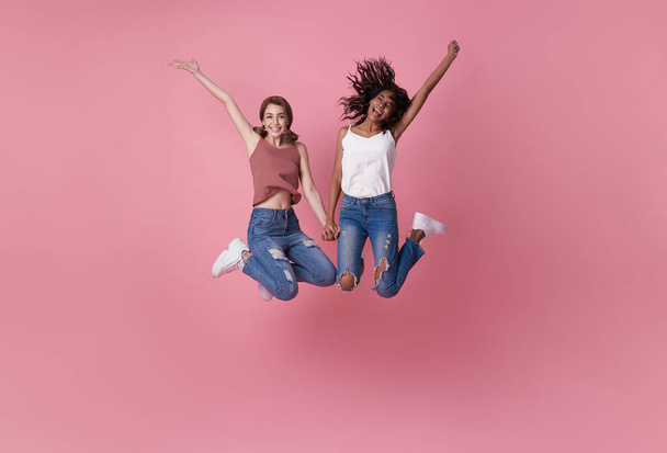 Δύο χαρούμενες ανέμελες νεαρές γυναίκες που πηδάνε στον αέρα με υψωμένες γροθιές κοιτάζοντας την κάμερα απομονωμένη σε ροζ φόντο.  - Φωτογραφία, εικόνα