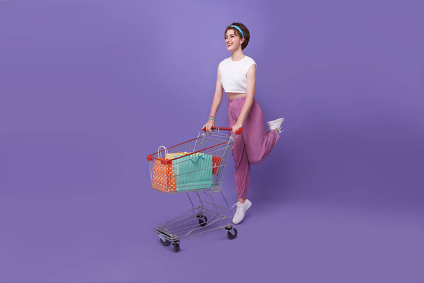 Cheerful young happy woman pushing shopping cart with shopping bag inside on purple studio background. - Photo, Image