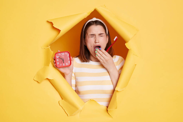 Portrait of sleepy woman wearing striped shirt and hair band, breaks through yellow paper background, holding alarm clock and toothbrush, yawning, covering mouth with palm. - Fotó, kép