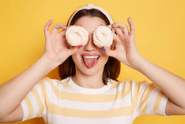 Playful childish woman holds marshmallows in hands, wearing striped shirt and glasses, covering her eyes with sugary desert, showing tongue out, having fun, posing isolated over yellow background. - Zdjęcie, obraz