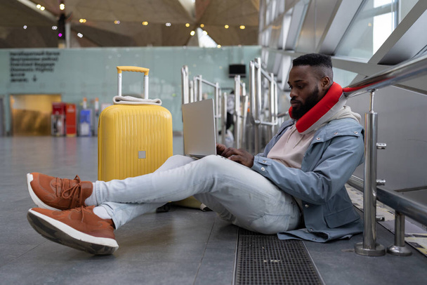 濃縮された若い黒人男性は、ソーシャルメディアで通信する黄色のスーツケースと首のフライト枕を身に着けているラップトップを使用して飛行を待っている足を組んで座って、情報をオンラインで検索し、電子メールを入力します - 写真・画像