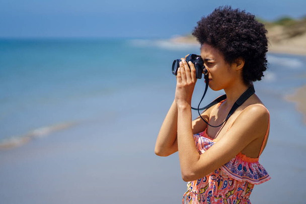 Молодая черная женщина с афроволосами в летнем платье, фотографирующая на пляже SLR-камерой. Девушка наслаждается отдыхом в прибрежной зоне. Свобода - Фото, изображение