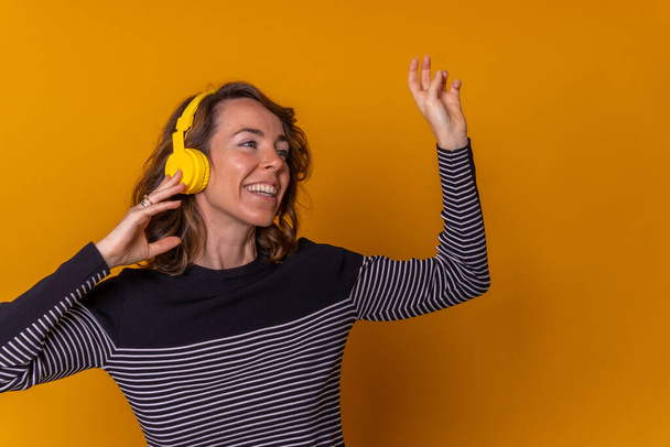Caucasian girl listening to streaming music and smiling and dancing with her phone, disco concept, headphones, yellow background - Foto, imagen
