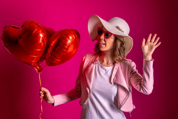 Portrait of a caucasian woman enjoying dancing with a white hat in a nightclub with some heart balloons, pink background - Fotografie, Obrázek