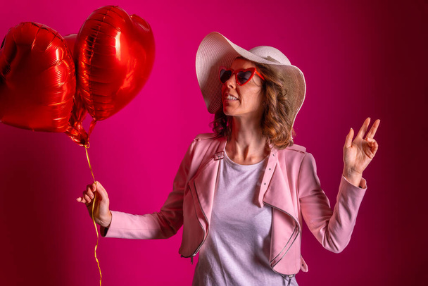 Portrait of a caucasian woman enjoying dancing with a white hat in a nightclub with some heart balloons, pink background - Φωτογραφία, εικόνα