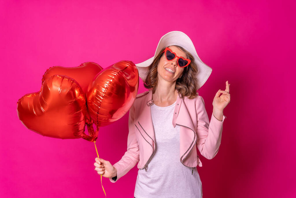 Portrait of a caucasian woman having fun with a white hat in a nightclub with some heart balloons, pink background - Photo, Image