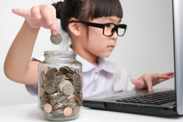 Mała bizneswoman wkłada monetę do szklanego słoika na stole, pracując z laptopem w biurze. Koncepcja dzieci i biznesu - Zdjęcie, obraz