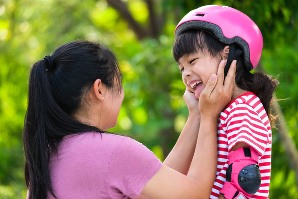 Madre asiática ayuda a su hija a ponerse almohadillas protectoras y casco de seguridad antes de practicar patinaje sobre ruedas en el parque. Emocionantes actividades al aire libre para niños. - Foto, Imagen
