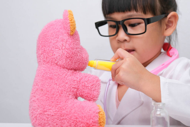 Cute Asian little girl playing doctor role game is giving injection to her sick teddy bear friend. Homeschool children's play and learning. - Photo, Image