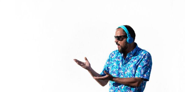 young hipster man with a beard, sunglasses, hawaiian shirt and blue headphones makes a surprised face showing what is next to him with both hands. Surprise face. copy space. White background - Photo, Image