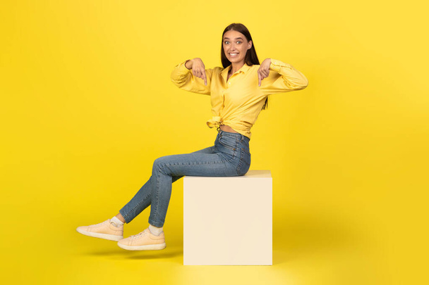 Look Here, Check This. Portrait of excited young woman pointing two fingers down at free copy space sitting on big white cube over yellow orange wall. Positive female showing empty mockup place for ad - Photo, Image