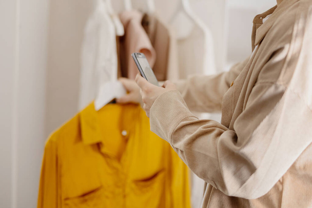 Frau hält Smartphone in der Hand und fotografiert ihre alten Kleider, um sie online zu verkaufen. Verkauf auf der Website, E-Commerce. Wiederverwendung, Second-Hand-Konzept. Bewusster Verbraucher, nachhaltiger Lebensstil. Nahaufnahme - Foto, Bild