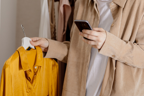 Женщина держит смартфон и фотографирует свою старую одежду, чтобы продать ее онлайн. Продажа на сайте, электронная коммерция. Повторное использование, подержанная концепция. Сознательный потребитель, устойчивый образ жизни. Крупный план - Фото, изображение