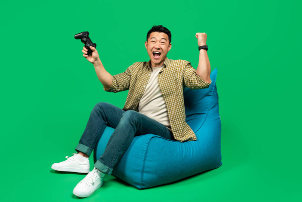 Joueur masculin mature heureux avec joystick faisant geste OUI, se sentant heureux sur la victoire, assis dans une chaise de haricot sur fond de studio vert - Photo, image