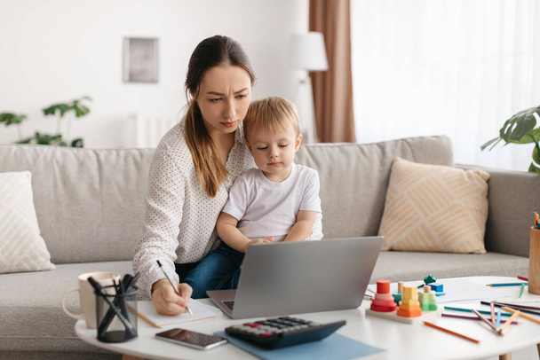 Junge Mutter Multitasking, Arbeit auf dem Laptop, während sie sich um ihren kleinen Sohn kümmert, Dame im Mutterschaftsurlaub, die versucht, sich selbstständig zu machen, mit Kind auf der Hand auf dem Sofa sitzend, Kopierraum - Foto, Bild