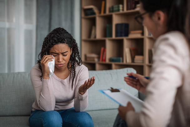 Депрессивная молодая черная леди плачет на консультации с европейской женщиной-психологом с планшетом, жестом в интерьере клиники. Психиатрическая помощь, помощь в решении проблем, терапия и медицинская поддержка - Фото, изображение