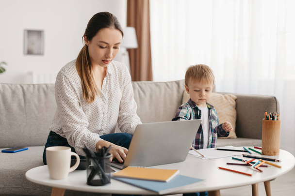 Freiberuflichkeit und Mutterschaftsurlaub. Junge Mutter arbeitet von zu Hause aus am Laptop und kümmert sich um ihren kleinen Sohn, Frau benutzt PC, während Kind in der Nähe spielt - Foto, Bild
