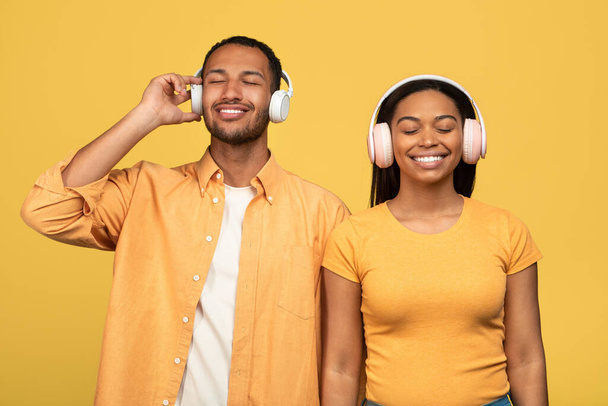 Веселая молодая чёрная пара слушает музыку в беспроводных наушниках на жёлтом фоне студии. Счастливые афроамериканские супруги отдыхают и наслаждаются аудиокнигой или любимыми песнями - Фото, изображение
