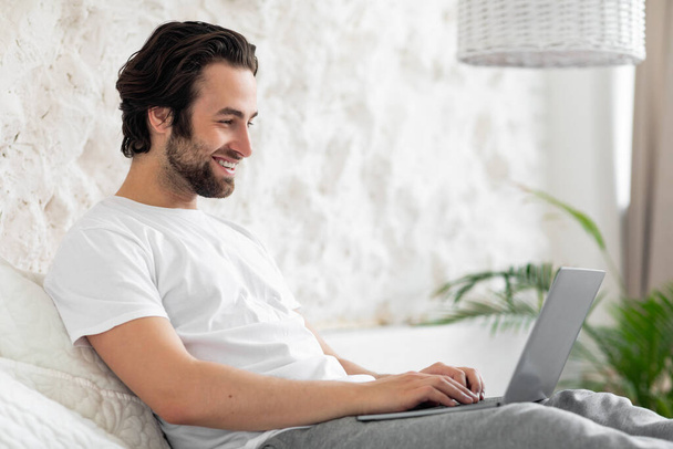 Positiv aussehender junger Mann, der zu Hause im Bett sitzt, online am Laptop arbeitet, auf der Computertastatur tippt, Freiberufler mit Kunden chattet, Freiraum, Schlafzimmereinrichtung. Entferntes Beschäftigungskonzept - Foto, Bild