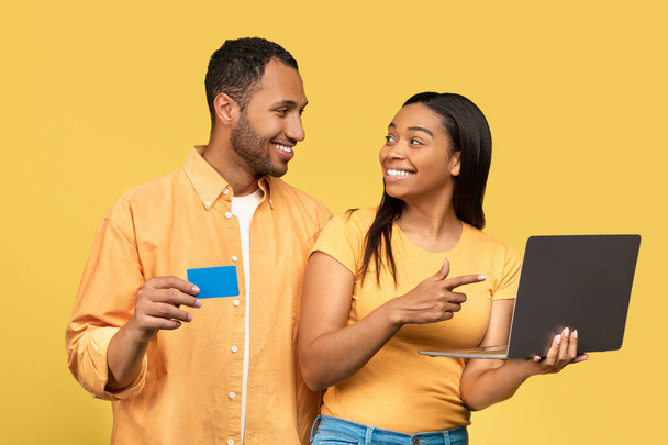 Portrait de famille millénaire noire tenant ordinateur portable et carte de crédit pour les achats en ligne sur fond de studio jaune. Joyeux couple afro-américain utilisant l'argent électronique pour acheter des marchandises sur le web - Photo, image