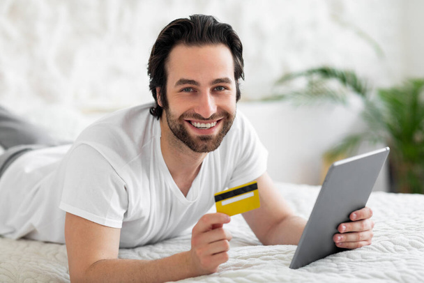 Cheerful young man using brand new digital tablet and plastic credit card while laying in bed at home, shopping on Internet, making online order while staying home, closeup portrait, copy space - Фото, зображення