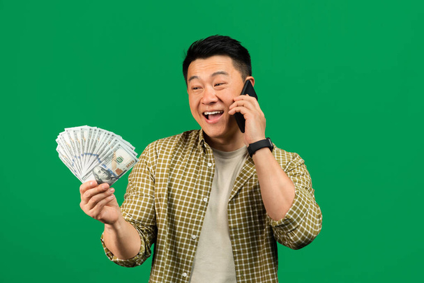 Alegre hombre asiático maduro sosteniendo dinero en efectivo y hablando en el teléfono inteligente, hombre afortunado reaccionando emocionalmente a ganar en línea, de pie sobre el fondo verde, espacio de copia - Foto, imagen