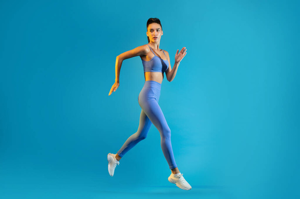 Fitness Female Jumping Exercising Posing In Mid Air Looking At Camera Over Blue Studio Background. Sportswoman Running Wearing Fitwear. Sport And Workout Concept. Full Length - Foto, Imagen
