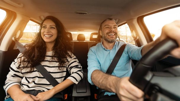 Happy man and woman going on summer vacation by car. Beautiful couple sitting in their new auto, cheerfully smiling looking at road, making test drive in the city, sun flare, windshield view - Foto, Imagen